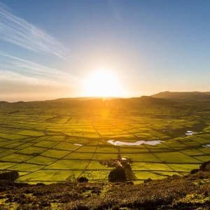 Il sole tramonta su un vasto paesaggio di campi verdi, tingendo le colline lontane di un caldo bagliore dorato sotto un cielo azzurro.