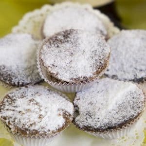 Un primo piano di alcuni piccoli dolci rotondi in pirottini di carta, abbondantemente cosparsi di zucchero a velo e disposti su un centrino di carta bianco.