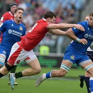 Un giocatore di rugby in maglia blu corre con la palla mentre un avversario in maglia rossa tenta di placcarlo.