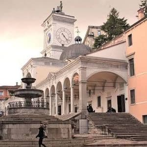 Una persona passa davanti a una grande fontana in pietra in una piazza davanti a un edificio classico con una torre dell'orologio, una cupola e un portico ad archi.