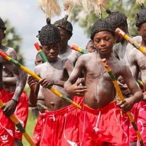 Un gruppo di giovani ragazzi in abiti tradizionali con capelli intrecciati e pittura corporale tiene bastoni colorati durante una cerimonia.