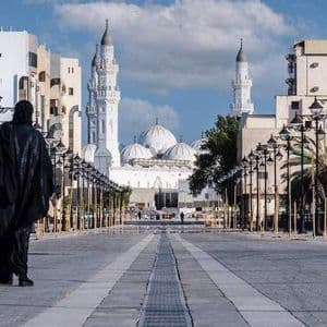 Una persona che indossa un'abaya nera cammina lungo un ampio lungomare fiancheggiato da lampioni decorati verso una grande moschea bianca con minareti.