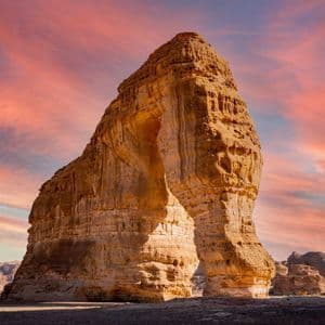 Un'imponente formazione ad arco di arenaria si erge in un paesaggio desertico sotto un cielo rosa e arancione vibrante durante il tramonto.