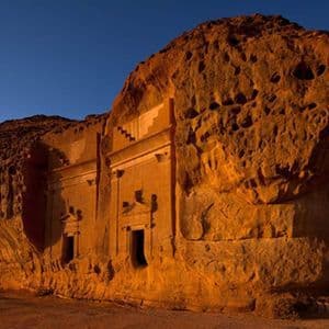 Una facciata di antica tomba scavata in una grande roccia arenaria, illuminata da una calda luce solare sotto un cielo azzurro e limpido.