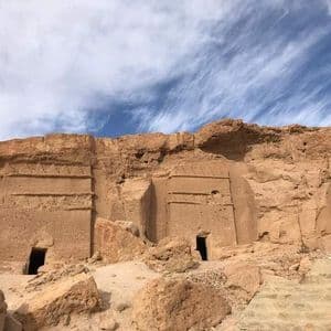 Antiche tombe con facciate rettangolari scavate in una scogliera rocciosa di arenaria sotto un cielo blu con nuvole sottili.