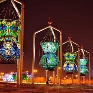 Una fila di grandi lanterne a mosaico colorate, appese a strutture e illuminate di notte, con lo skyline della città sullo sfondo.