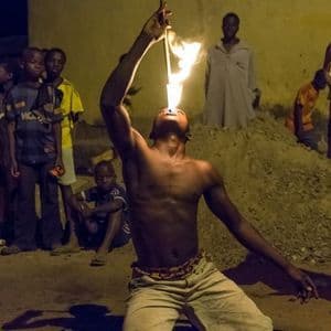 Un uomo a torso nudo si inginocchia a terra di notte, sputando fuoco da un bastone mentre una folla di persone guarda.