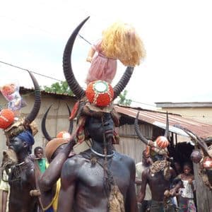Uomini con pittura corporea scura indossano elaborati copricapi cornuti, uno dei quali con una bambola, durante una cerimonia del villaggio circondati dalla folla.