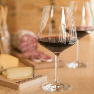 Un bicchiere di vino rosso poggia su un bancone di legno, con taglieri di salumi e formaggi sfocati sullo sfondo.