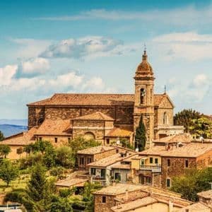 Una chiesa in pietra con un campanile domina un borgo collinare con tetti in terracotta, con le montagne sullo sfondo.