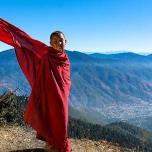 Un ragazzo in abiti rossi si erge su una cima di montagna, la sua veste che sventola al vento sopra una valle.