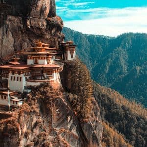 Ein Klosterkomplex mit weißen Mauern und roten Dächern schmiegt sich an eine steile Klippe und überblickt bewaldete Berge unter blauem Himmel.
