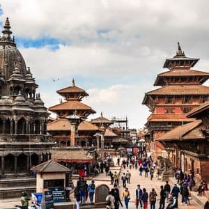 Ein belebter Stadtplatz voller Menschen, mit einem großen, verzierten Steintempel und traditionellen mehrstöckigen Gebäuden unter einem teilweise bewölkten Himmel.