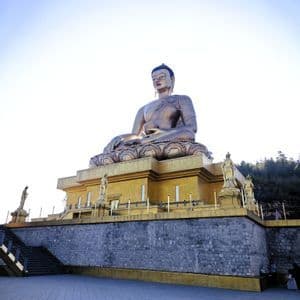 Eine riesige Bronzestatue eines sitzenden Buddhas auf einem goldenen Podest, von einem steinernen Innenhof unter hellem Himmel gesehen.