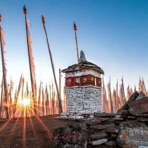Eine kleine steinerne Stupa thront auf einem felsigen Hügel, umgeben von weißen Gebetsfahnen, während die Sonne aufgeht und einen Sonnenstern am Horizont bildet.