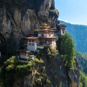 Ein Kloster mit weißen Wänden und verzierten goldenen Dächern ist in die Seite einer steilen, felsigen Klippe gebaut, die ein bewaldetes Tal überblickt.