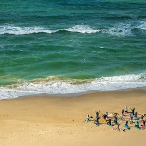 Un gruppo WeRoad visto dall'alto, che pratica yoga su tappetini colorati su una spiaggia di sabbia, accanto alle onde turchesi dell'oceano.