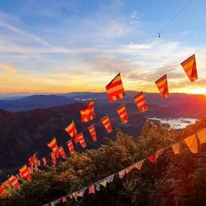 Bunte buddhistische Gebetsfahnen hängen über einem bewaldeten Bergtal mit einem Gewässer bei Sonnenaufgang.