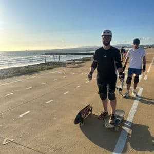 Due uomini di un viaggio di gruppo WeRoad, con caschi e protezioni, sono in piedi con i loro skateboard su un percorso costiero pavimentato al tramonto.