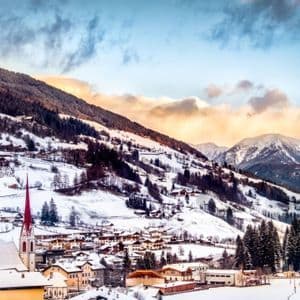 Un villaggio innevato con una chiesa dalla guglia rossa si adagia ai piedi di una catena montuosa boscosa, sotto un cielo nuvoloso al tramonto.