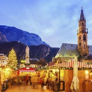 Un mercatino di Natale splendidamente illuminato in una piazza cittadina al tramonto, con una cattedrale storica e montagne sullo sfondo.