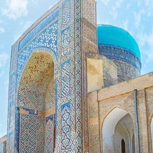 Froschperspektive auf ein historisches Gebäude mit einem Bogentor, aufwendigen blauen Mosaiken und einer großen türkisen Kuppel vor einem teils bewölkten Himmel.