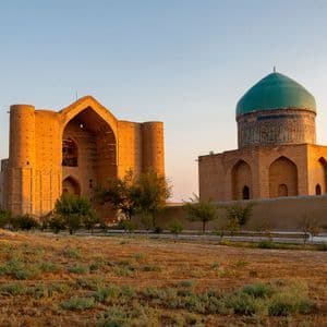 Two historic brick buildings, one with a large turquoise dome, illuminated by golden hour light across a grassy field.