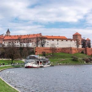 Un imponente castello storico su una collina erbosa, che domina un fiume, con persone che passeggiano lungo la riva e una barca ormeggiata sull'acqua.