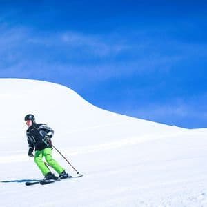 Eine Person mit Helm und grüner Hose fährt einen sonnenbeschienenen, schneebedeckten Hang unter blauem Himmel hinunter.