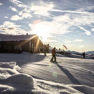 Eine Person in roter Jacke fährt bei Sonnenuntergang auf einem verschneiten Berghang vor einer Hütte Ski.