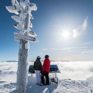 Zwei Skifahrer stehen auf einem verschneiten Berggipfel und blicken über ein Wolkenmeer unter strahlender Sonne neben einem gefrorenen Wegweiser.
