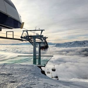 Eine WeRoad Gruppenreise im Skilift, der von einer verschneiten Bergstation über einem Wolkenmeer abfährt.