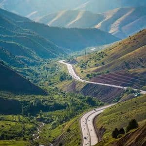 Veduta aerea di una strada serpeggiante con auto che percorrono una vasta e verde valle montana sotto un sole velato.