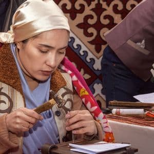 Una donna con un foulard si concentra mentre modella un pezzo di metallo con un piccolo martello su un tavolo da artigianato.