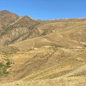 Una persona con una maglietta blu e un berretto bianco si trova su un'ampia collina arida con aspre montagne sullo sfondo, sotto un cielo azzurro e limpido.