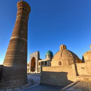 Un alto minareto in mattoni decorati e edifici a cupola si ergono in un cortile lastricato sotto un cielo limpido e azzurro.