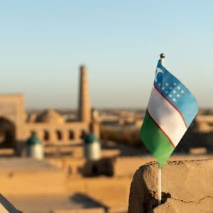 Primo piano della bandiera dell'Uzbekistan posizionata su una superficie di pietra, con uno sfondo sfocato dello skyline di una città antica al tramonto.