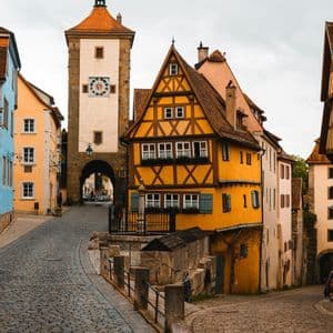 Una via acciottolata e tortuosa costeggia edifici colorati a graticcio e una torre dell'orologio in pietra in un centro storico.