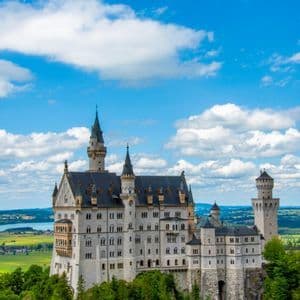Un grande castello bianco con molte torri si erge su una collina lussureggiante e boscosa sotto un cielo blu con nuvole bianche.