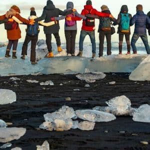 A WeRoad group trip with their arms around each other, standing on a large block of ice on a black sand beach.