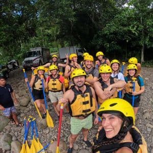 Un gruppo WeRoad con caschi gialli e giubbotti di salvataggio posa per un selfie sulla riva rocciosa di un fiume prima di fare rafting.