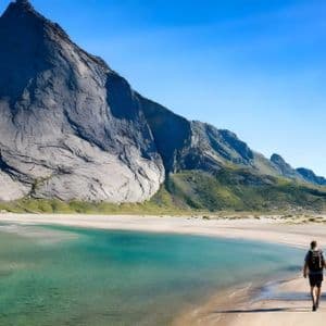 Due escursionisti con zaini camminano su una spiaggia sabbiosa accanto all'acqua turchese ai piedi di un'imponente montagna rocciosa.