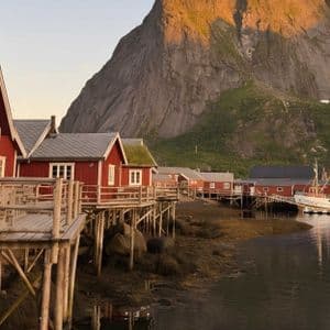 Case di legno rosse su palafitte si allineano lungo una tranquilla costa, con un'ampia montagna rocciosa sullo sfondo al tramonto.