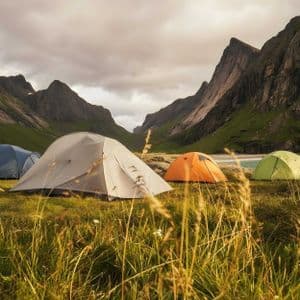 Vier farbige Zelte stehen auf einer grünen Wiese am Fuße großer, felsiger Berge neben einem Gewässer.