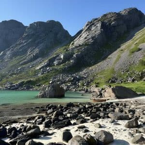 Ein felsiger Strand mit einem kleinen Holzboot trifft auf ruhiges türkisfarbenes Wasser am Fuße steiler Berge unter einem klaren blauen Himmel.