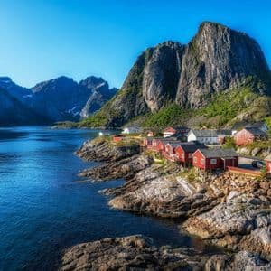 Ein Dorf mit roten Holzhäusern, an einer felsigen Küste neben einem blauen Fjord gebaut, mit steilen Bergen im Hintergrund.