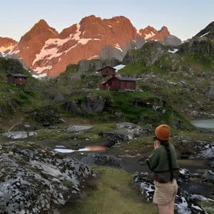 Eine Frau mit gelber Mütze steht auf einem Pfad mit Blick auf ein grünes Tal mit roten Hütten, einem See und sonnenbeschienenen, schneebedeckten Berggipfeln.