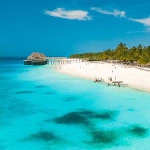 Vista aerea di una spiaggia di sabbia bianca con acqua turchese, palme e un pontile di legno con una palafitta sotto un cielo blu.