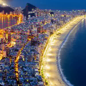 Vista aérea nocturna de una ciudad costera brillantemente iluminada con una playa larga e iluminada que se curva a lo largo del oscuro océano.