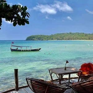 Une table et des chaises sur une terrasse en bois surplombent une mer turquoise avec un bateau ancré et une plage tropicale lointaine.